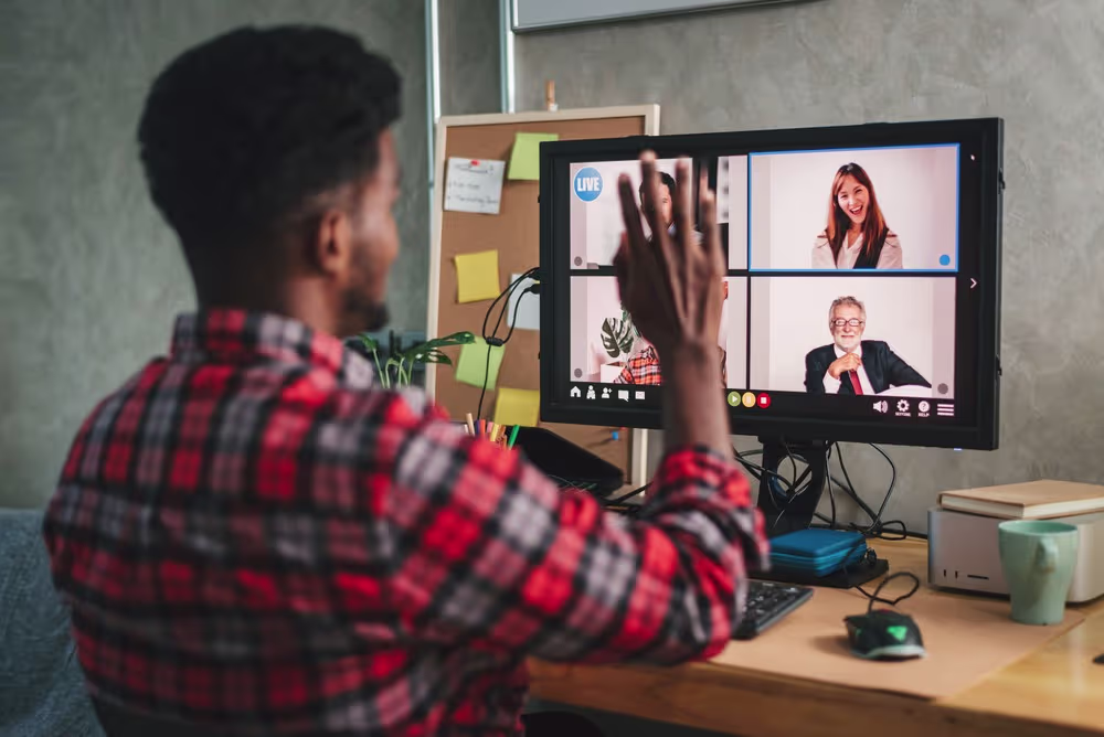 A man is waving on a live video call with colleagues shown on a monitor at his desk.