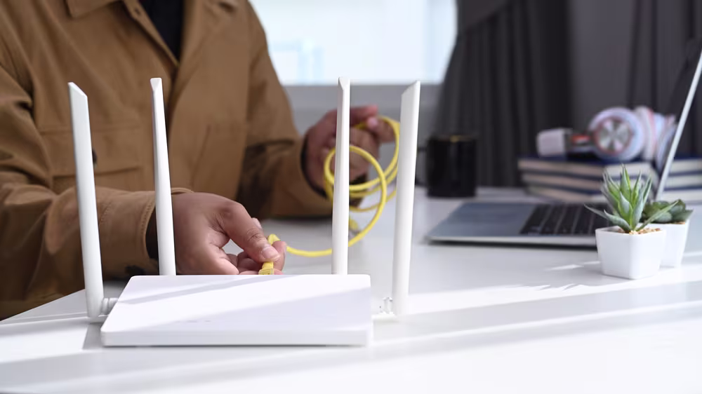 A person connects a yellow Ethernet cable to a Wi-Fi router on a desk, illustrating slow network performance