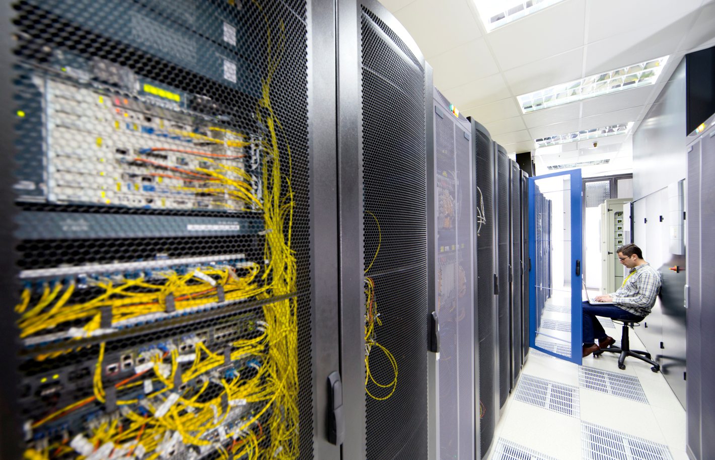 A technician working on a laptop between rows of server racks in a large data centre, illustrating bandwidth bottlenecks